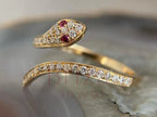 A gold snake ring with clear pavé diamonds and red gemstone eyes, displayed on a textured gray stone surface.