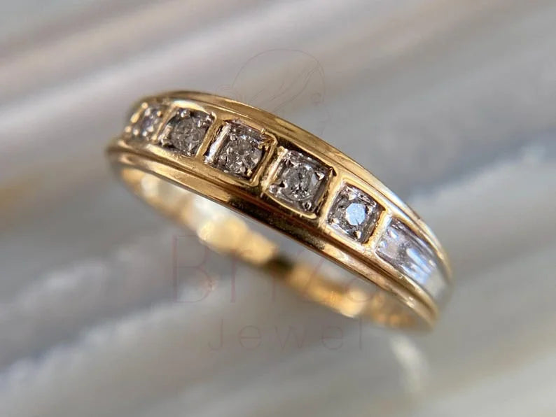 A yellow gold eternity ring featuring a row of square-cut clear gemstones and channel-set round stones, shown close-up on a light marble surface.