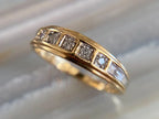 A yellow gold eternity ring featuring a row of square-cut clear gemstones and channel-set round stones, shown close-up on a light marble surface.